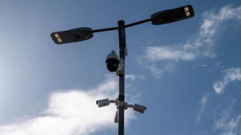 Equipment on a black pole including three ANPR cameras, a CCTV camera and lights. The sky is the backdrop.