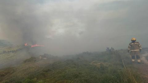A firefighter stands on a track in a smoke-filled landscape with a fire burning in the distance.