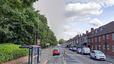 A bus stop is on the left of the road in front of a row of trees with houses on the other side and cars parked outside.