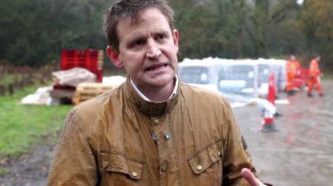 A man in a brown coat standing in a car park.