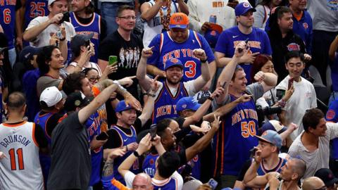 Rows of people mostly wearing jerseys in the Knicks colors of orange and blue throw their hands in the air, cheering a victory.