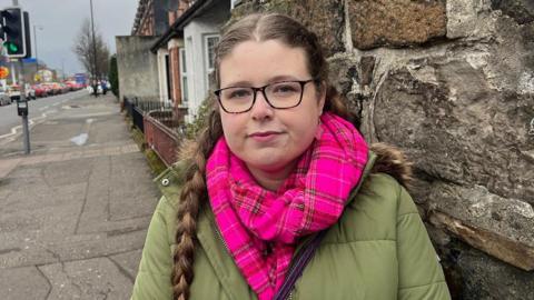 Historian Dr Robyn Atcheson - She has her hair in two braids and is wearing a pair of square-framed glasses, a pink plaid scarf and a green puffer jacket. She is standing on the side of the street near a stone wall.