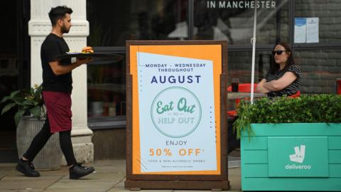 A waiter carries a tray with a burger on it to a lone diner who sits under and umbrella in the shade. She has sunglasses on and her arms folded