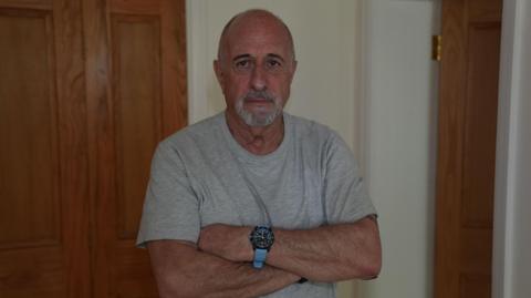 A man with closely cropped grey hair and a grey goatee beard wears a grey t-shirt and a black and light blue wristwatch. His arms are folded in front of him as he stands in front of two wooden doors.