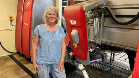 A middle-aged woman with mid-length blonde curly hair, a pale blue top and light blue, dirty jeans stands in front of a red and grey robotic device on a farm