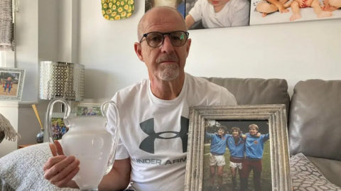 A man in a white t-shirt, who is bald and wears glasses, sits on a sofa in a living room, holding a replica of a trophy and a photo frame.
