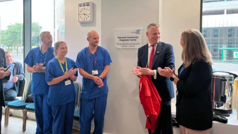 Health staff and MP Luke Pollard stand inside the new centre next to a plaque. There are large windows behind them. The group of people are clapping their hands together and are smiling at one another.