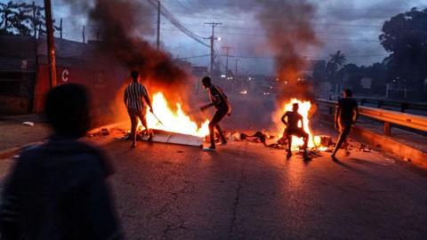 Four silhouetted individuals stand near burning barricades in the middle of a road during an evening protest in Maputo, Mozambique. Thick smoke rises into the dark, cloudy sky. Faint lights and palm trees are visible in the background.