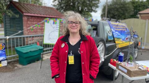A woman in a red coat is standing in a car park next to a model of a pylon and in front of a car draped in protest banners