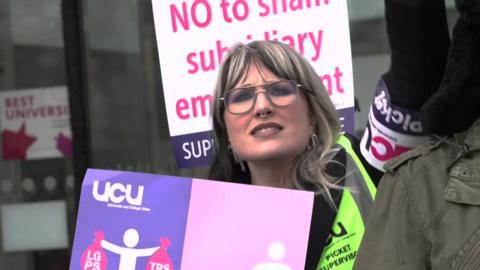 A woman with long blonde hair has one arm in the air. She is holding a sign that says no to subsidiary employment. She is wearing a UCU armband.