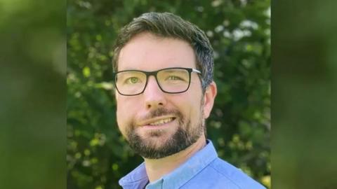 Aaron Jones looking at the camera. He is wearing a blue shirt and dark-rimmed glasses and dark hair. He has a short trimmed beard and moustache