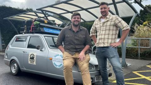 Two men are standing in front of a grey car. The car has stickers on the side and luggage strapped to the roof.