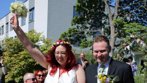 A woman wearing a wedding dress with flowers in her hair and a man wearing a black wedding jacket over a white shirt and green tie. The woman is holding aloft a bouquet of flowers. They are both smiling and other men and women - or wedding guests - can be seen behind them. They are all outside. 