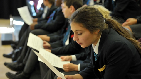 School pupils sat in a line with school girl in foreground reading.