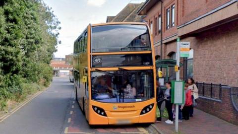 Passengers board a bus at Maylord Orchards in Hereford. The bus is orange and the bus shelter has a green frame and stands next to the bus stop sign. There is a marked bus stop bay in the road, which has a building on one side and trees on the other.