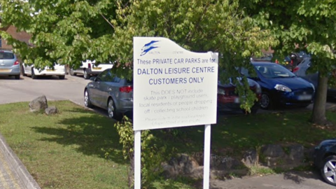 A sign on a grassy verge in a car park saying Dalton Leisure Centre Customers Only. There are some cars in the background.
