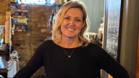 A blonde woman smiling at the camera. She is standing behind the bar in a pub. She is wearing a black top with a gold necklace.