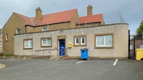 In the back ground is a two-storey building. In the foreground is a single-storey grey building with flat roof.
