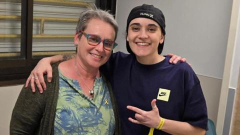 Emily Damari (R) stands smiling with her right arm around her mother Mandy's shoulders while holding up her left hand with two fingers missing