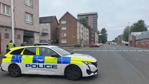 Police car with police tape closing off a street in Paisley. Blocks on flats line the pavements and police officers stand on the edge of the cordon.