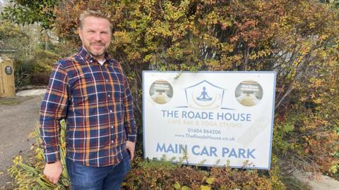 A man in a navy, red, yellow and yellow striped shirt and jeans stands next to a white sign for The Roade House which is the the middle of a hedge. 