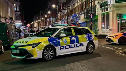 A night-time shot of a police car foreground on a street with police tape and officers behind 