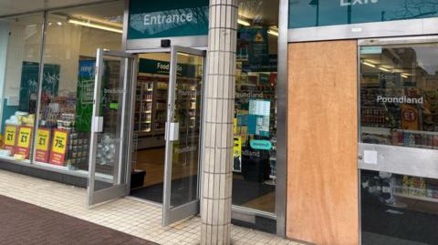 A damaged Poundland shopfront. There is a glass door boarded up and grey graffiti on the glass.