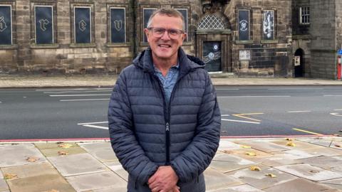A man with short grey hair, glasses and wearing a dark blue coat over a blue shirt, stands and smiles with his hands clasped in front of him. Behind him is a road and an old building on the other side.