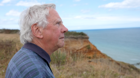 Derrick Bowler, a resident in Trimingham in north Norfolk who is standing on a cliff edge. He is not looking directly at the camera and is instead looking out to sea, which is to the right of the photograph. He has grey white hair and is wearing a blue checked shirt.