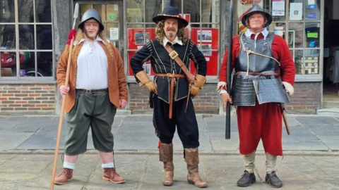 Three men - all in historical costumes dating from the English Civil War era - standing in front of an ancient-looking shop in Gloucester