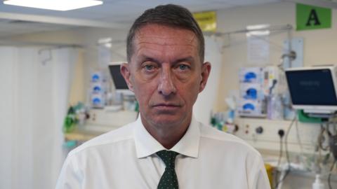 A middle-aged man faces the camera wearing a white shirt and black tie with the equipment of a hospital ward seen behind him