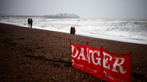 Danger sign on a beach warning of rough seas.  Two couples are looking out to the sea with a damaged pier in the background.