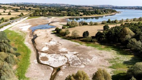 A drone shot of Chew Valley Lake showing the water line receding around 100 metres from its usual line and exposing dry mud. It is a bright sunny day and the grass in the fields is yellowed
