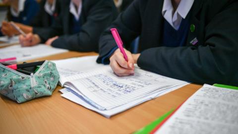 Close-up of unidentified girls in uniforms writing on notebooks on their school desk. There is a green pencil case on the left with lined books full of notes around. A girl writes with a pink pen.