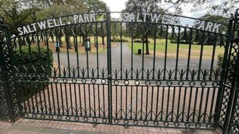 A large and ornate pair of black gates with the words "Saltwell Park" written in white letter across the top of each. They are closed and beyond them is a park with large green areas divided by footpaths with trees and bushes.
