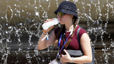 A woman wearing sunglasses and a black sun hat drinking a bottle of water. She is wearing a red t-shirt and surrounded by water droplets from a fountain