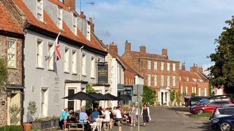 The view from the centre of Burnham Market. There are people siting at picnic tables outside a pub.