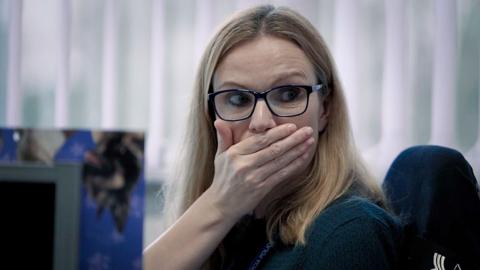 A woman with long blonde hair and glasses puts her hand over her mouth in surprise