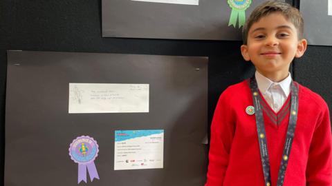 Five year old boy in a red school jumper smiles with his winning engineering design