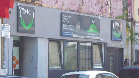 Exterior of a grey building with black doors and windows and peeling paint on the first storey. Above the entrance is a sign saying "IMC" for Inverclyde Muslim Centre.