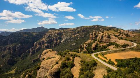 An image depicting the hills of Tremp in Catalonia. Large green and brown mountains stand in front of a blue sky with several clouds. There is a winding road through the mountains on the left of the image 