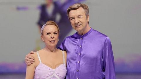 Sir Christopher Dean stands with his skating partner Dame Jayne Torvill in front of a mural showing them skating, both are wearing purple outfits