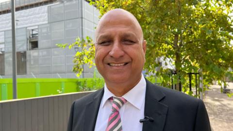 Naeem Ahktar wears a grey suit jacket and pink striped tie with a white shirt. He smiles at the camera.