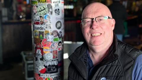 A bald man with black glasses is smiling. He is wearing a blue top with a black gilet jacket over it. Next to him is a supporting beam that is metal and covered in stickers