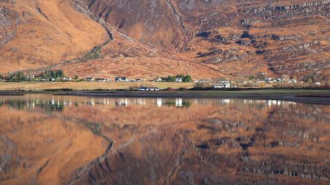 Torridon village and the mountainside that rises behind it. The village and mountain are reflected in the mirror-like waters of Loch Torridon.