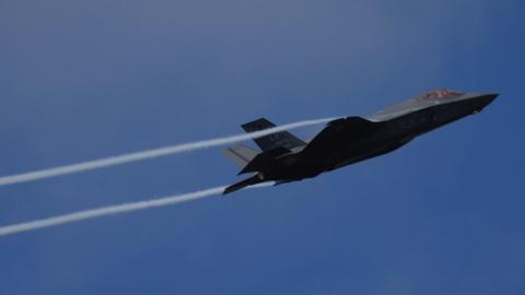 A USAF F-35A Lightning II, a grey military aircraft, flies left to right against a cloudless blue sky with two white vapour trails coming off its wings 