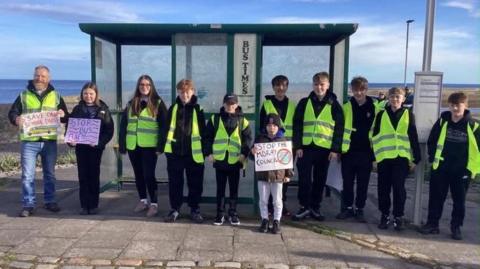 Campaigners with save school bus signs at a bus stop, wearing yellow hi-vis tops.