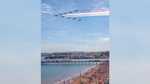 Nine Red Arrow aeroplanes fly in an aerial display in the sky over Torbay. There is a sandy beach with people on it and blue sea. There is a pier and houses in the distance.