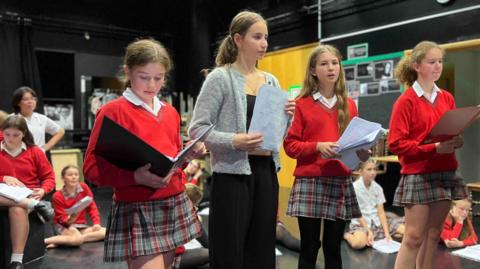 A group of four children standing in a rehearsal space, holding scripts. Three are wearing red sweaters and plaid skirts, while one is wearing black trousers and a grey cardigan. Several others are seated on the floor in the background.