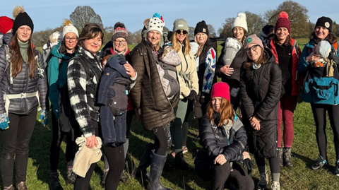 A group of women with their babies standing in a field. The sky is blue and they are all wrapped up for winter. They are smiling towards the camera. 